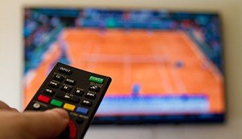 An up-close image of a person with their hand on a remote and a TV on in the background